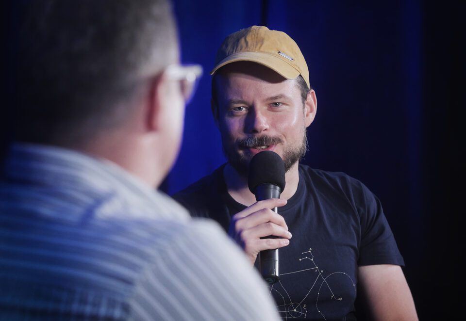 Zbliżenie na Wojtka Szota, zdjęcie zrobione podczas rozmowy; Szot przy ustach ma mikrofon, patrzy na rozmówcę i lekko się uśmiecha; na głowie ma brązowawą kaszkietówkę.