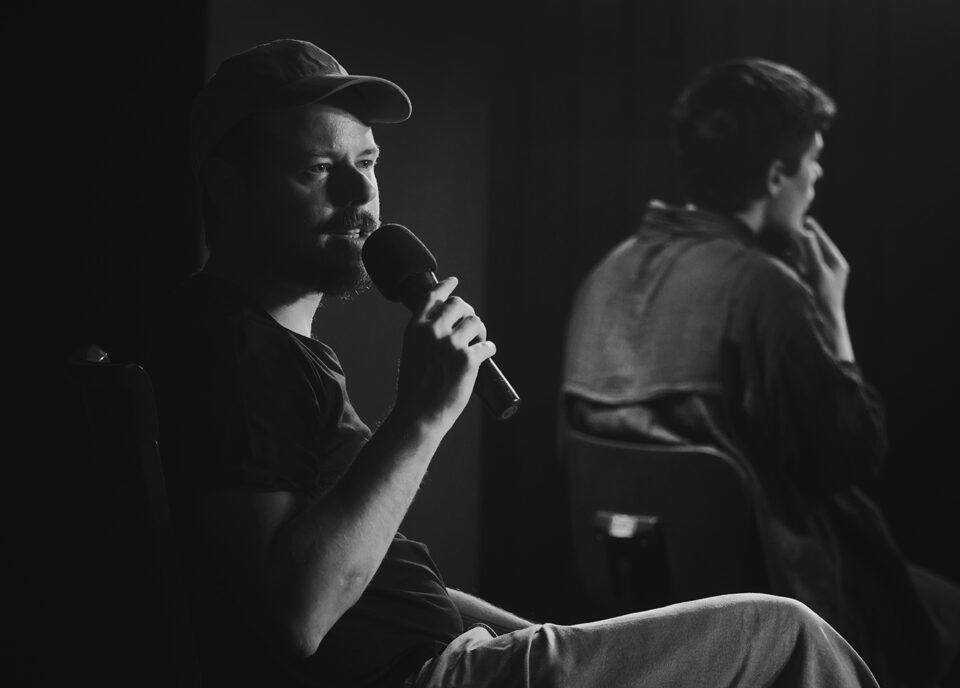 Czarno-biały portret Wojtka Szota; mężczyzna siedzi swobodnie na krześle, w ręku ma mikrofon; za nim tłumaczka języka migowego.