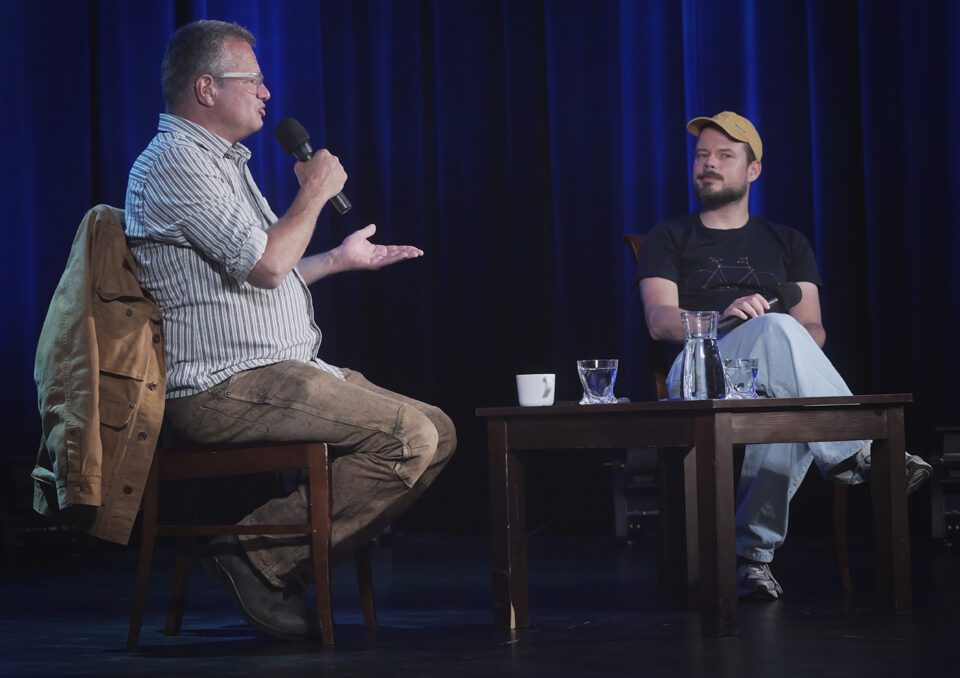 Meller i Szot podczas rozmowy; mężczyźni siedzą na krzesłach, Meller gestykuluje a Szot zerka poza kadr; pomiędzy nimi prosty stół, w tle podświetlona na niebiesko kotara sceny.