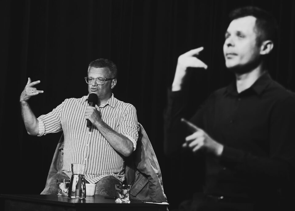 Zbliżenie na Mellera, mężczyzna gestykuluje, prawdopodobnie coś wylicza - ma wyciągniętą przed siebie dłoń z rozprostowaną częścią palców; podobny gest wykonuje tłumacz PJM - ten jest na 1. planie ale lekko rozmyty (nieostry).