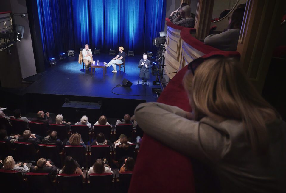 Zdjęcie zrobione z loży na widowni; na 1. planie kobieta, która opiera się o barierkę loży; w tle scena podczas rozmowy.