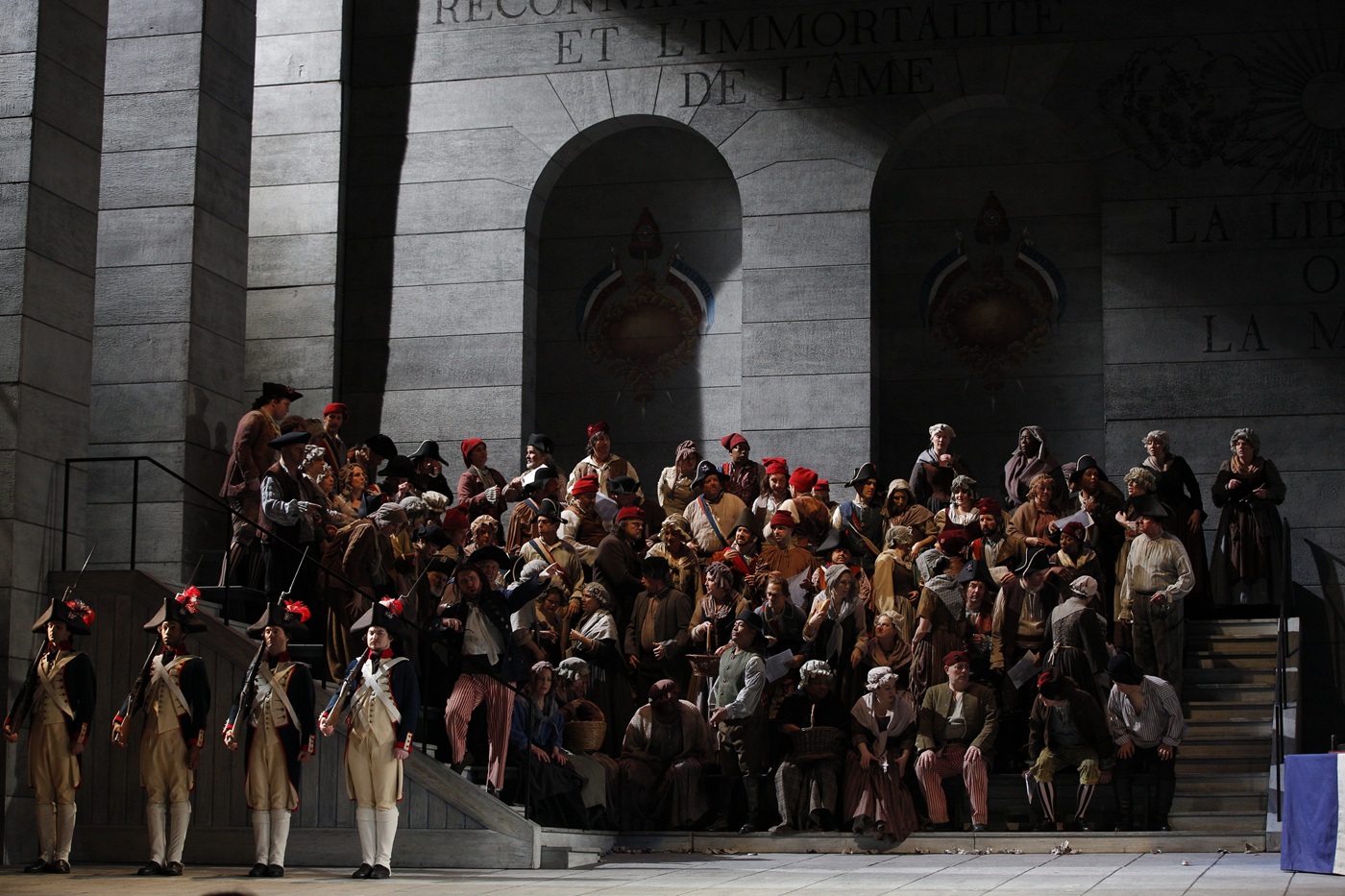 Tłum ludzi pod budynkiem. Ludzie są w strojach barokowych. Po lewej stronie schody, w ujęciu z boku, na schodach również tłoczy się tłum. Z tłumu wyróżniają się czterej strażnicy stojący przed ujętymi z boku schodami. Stoją na baczność w równych odstępach. Mają jasne stroje, ciemne surduty i czarne czapki z czerwonym pióropuszem. Każdy z nich trzyma strzelbę opartą na ramieniu. W tle fasada budynku z szarego kamienia z wysokimi wnękami zakończonymi półokrągło.