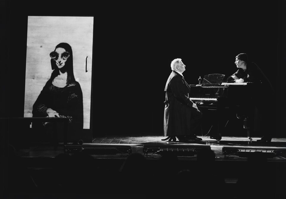 Czarno-białe zdjęcie sceny. Na scenie dwie osoby. Po prawej stronie kobieta i mężczyzna po przeciwnych stronach fortepianu. Mężczyzna w garniturze siedzi przy klawiaturze fortepianu. Po drugiej stronie wpatrzona w niego kobieta w czarnym stroju i czapce. Opiera ręce na klapie fortepianu. Po lewej stronie ekran z wyświetlonym kolażem z Mana Lisą z doklejonymi nienaturalnie wielkimi oczami. Tło czarne.