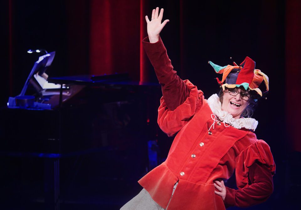 Kobieta w czerwonym stroju błazna, strój ma bufiaste rękawy, białą kryzę. Kobieta na głowie ma czapkę z trójkątnymi wykończeniami i dzwoneczkami; Kobieta wykonuje taniec - uśmiecha się szeroko, jest w lekkim rozkroku (ale nogi są poza kadrem), jej prawa dłoń jest wyciągnięta ku górze, lewą opiera na biodrze.