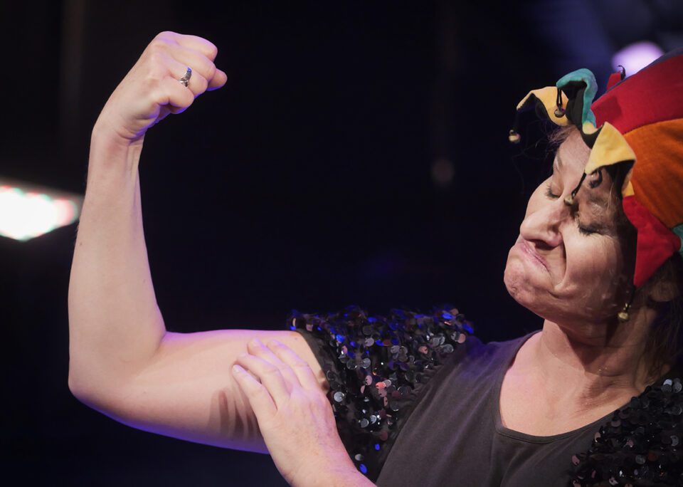 Zbliżenie, portret aktorki w czapce błazna, kobieta pokazuje biceps, napina mięśnie (są bardzo niewielkie) i robi zadziorną minę, ale ma przymknięte oczy.