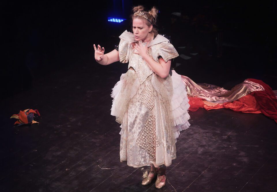 Kobieta stoi na scenie, prowadzi monolog i patrzy w stronę publiczności; jedną dłonią przykłada do ciała ozdobną, historyczną suknię; za nią na podłodze leży wiele czerwonych, różowych tkanin i czapka z trójkątów.