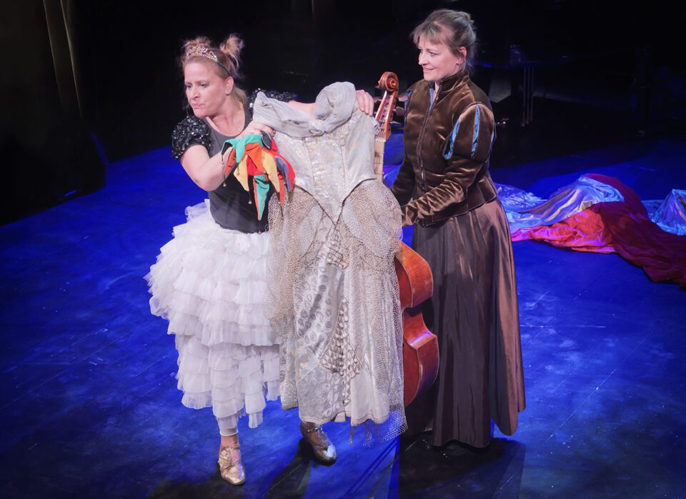 Duet - aktorka w tiulowej spódnicy i kontrabasistka, kobiety stoją obok siebie, kontrabasistka z instrumentem; aktorka trzyma w wyciągniętych dłoniach ozdobną, kremową suknię.