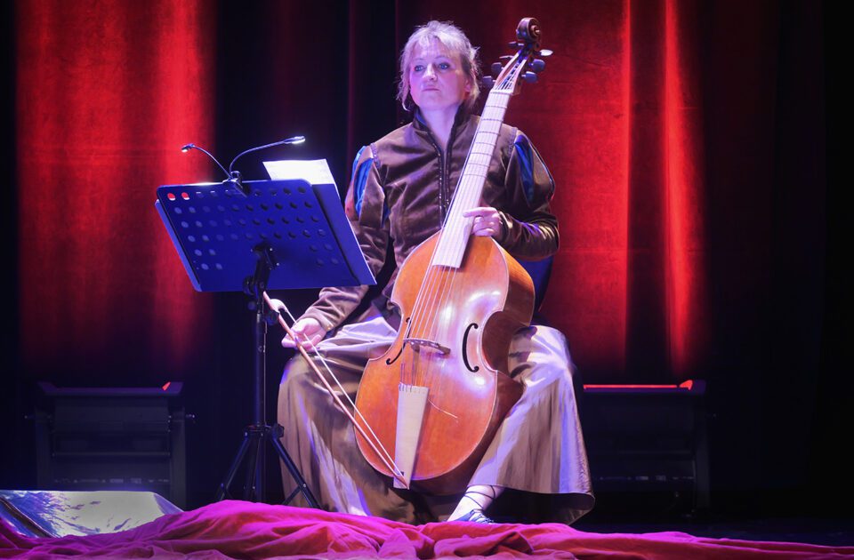 Zdjęcie kontrabasistki; kobieta zerka spokojnie na nuty na pulpicie przed nią. W tle czerwona kotara.