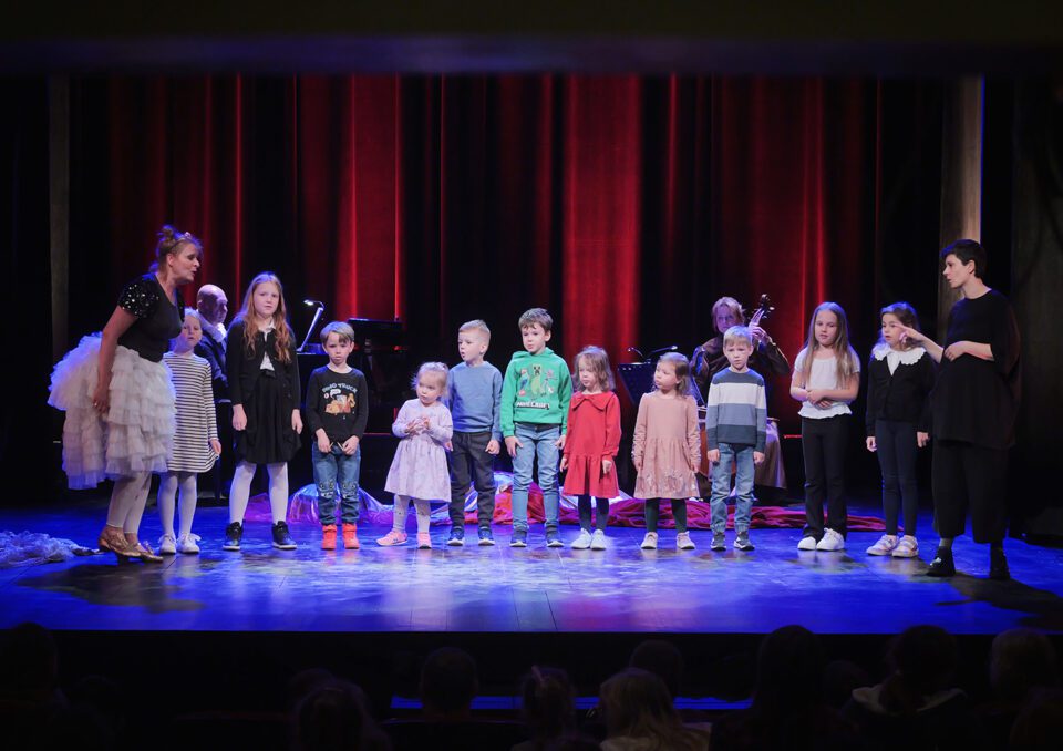 Grupa dzieci na scenie; stoją w rzędzie; po lewej stronie sceny aktorka w tiulowej spódnicy, po prawej - tłumaczka PJM; kobiety zachęcają dzieci do zabawy. Za dziećmi pianista i kontrabasistka. W tle czerwona kotara.