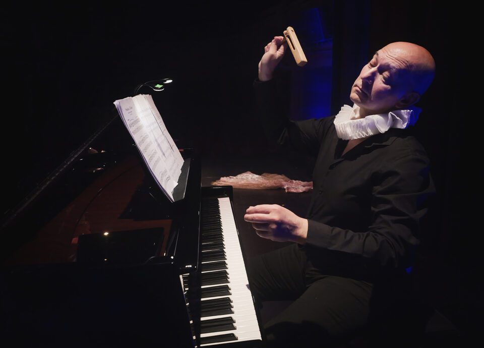 Pierwsze z cyklu 10 zdjęć; ujęcie na postać podczas gry na fortepianie. To łysy mężczyzna, który jest ubrany cały na czarno, na szyi ma białą kryzę - ozdobne drapowanie; mężczyzna ma w prawej dłoni drewnianą kołatkę.