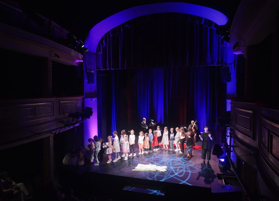 Widok na scenę Teatru Starego, na scenie dwoje muzyków - mężczyzna przy fortepianie i kobieta z kontrabasem, obok nich tłumaczka PJM; ze sceny schodzi właśnie grupa przedszkolaków.