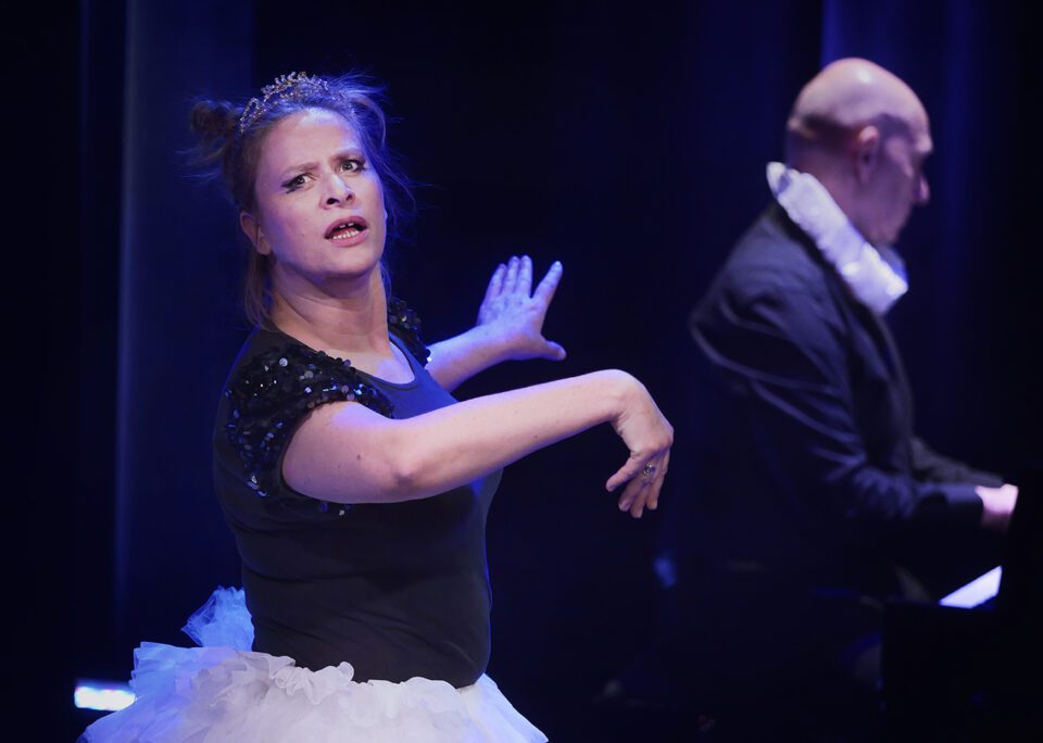 Na pierwszym planie jest kobieta, która ma na sobie tiulową spódnicę i bluzkę z rękawami obszytymi cekinami; na głowie ma diadem; kobieta wykonuje piruet; ma ręce wyciągnięte na boki; w tyle rozmyta sylwetka pianisty.