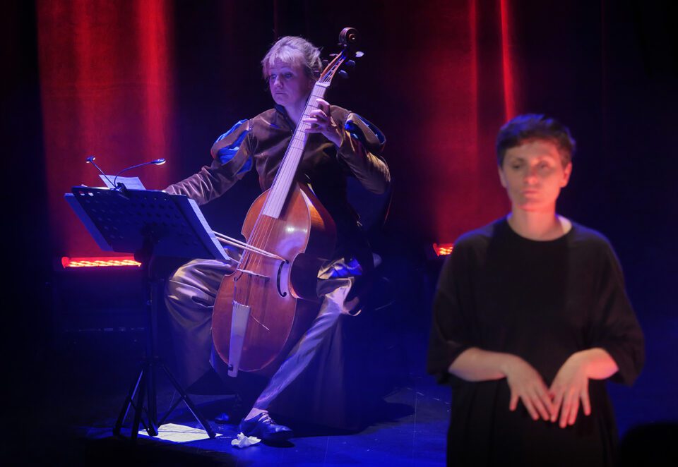 Portret kontrabasistki, kobieta ma na sobie ciemny połyskujący kostium z bufiastymi rękawami; gra na instrumencie; przed nią stoi pulpit z nutami; na pierwszym planie po prawej tłumaczka PJM.