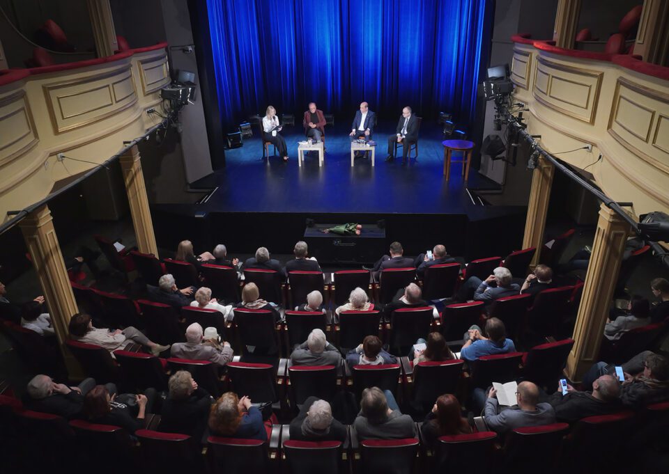 Zdjęcie wykonane z loży teatralnej na piętrze; na scenie rozmówcy; wszystkie miejsca na widowni zajęte.