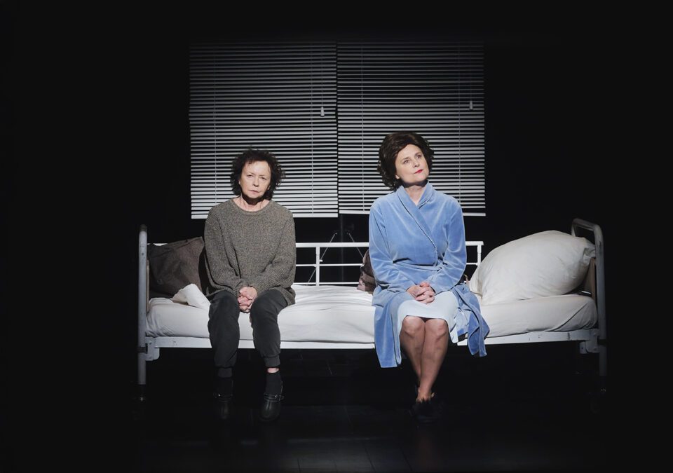 Zdjęcie dwóch kobiet siedzących na szpitalnym łóżku. Kobieta po lewej jest w czarnych spodniach i ciemnoszarym swetrze. Kobieta po prawej ma na sobie błękitny szlafrok. W tle ciemna ściana i jasne żaluzje.