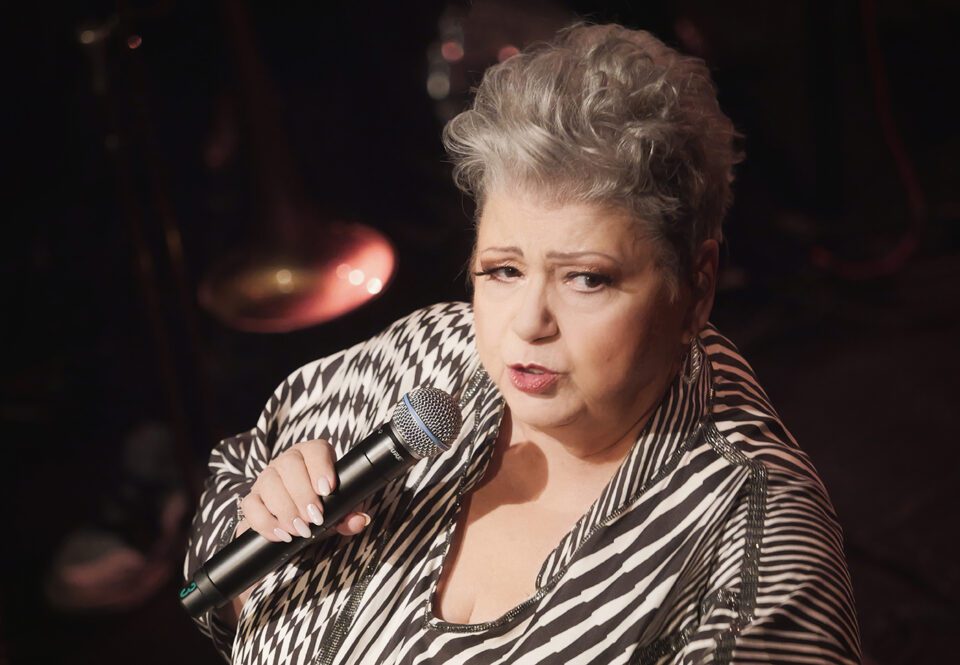 Portret Ewy Bem z mikrofonem. Kobieta ma krótkie kręcone, siwe włosy. Jest w stroju w pasy o różnej grubości. Tło ciemne.