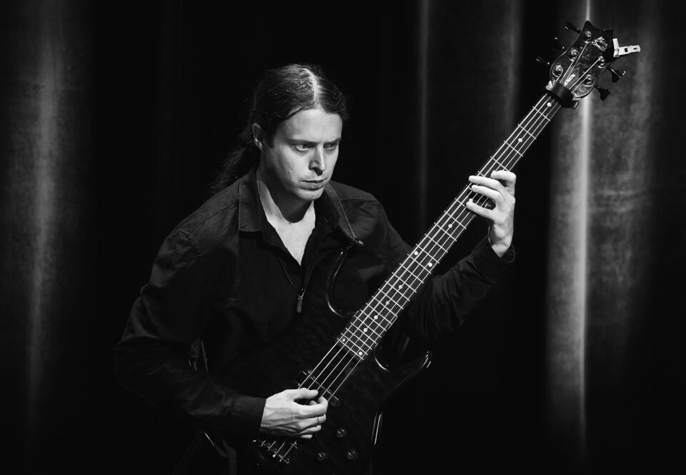 Czarno-białe zdjęcie mężczyzny grającego na gitarze elektrycznej. Mężczyzna jest w czarnej koszuli, ma długie włosy związane w kucyk. Tło ciemne.