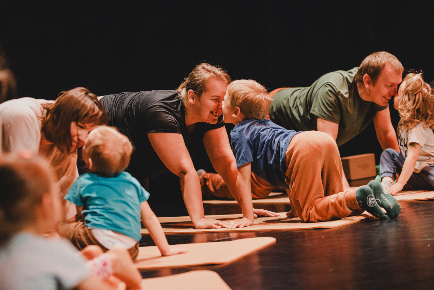 Zdjęcie dzieci z opiekunami. Wszyscy są uśmiechnięci w pozycji na czworaka. Każde z dzieci jest zwrócone w stronę swojego opiekuna. Tło ciemne.