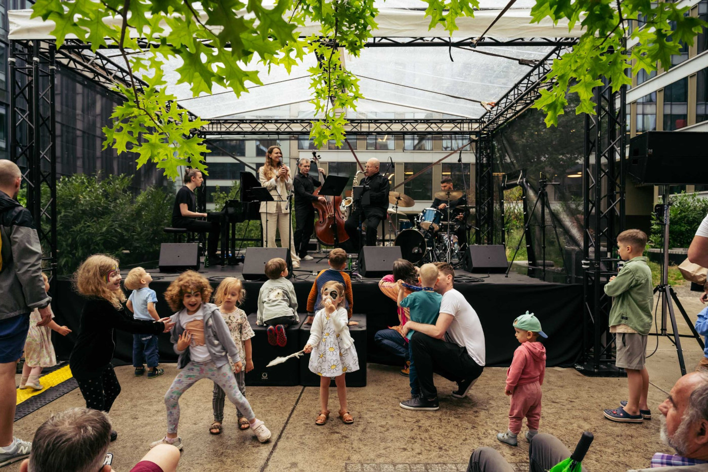 Pięcioosobowy zespół muzyczny na małej, scenie w plenerze. Scena jest czarna. Nad sceną jest przezroczysty dach na czarnej konstrukcji. Przed sceną jest grupa małych dzieci w ekspresyjnych pozach.