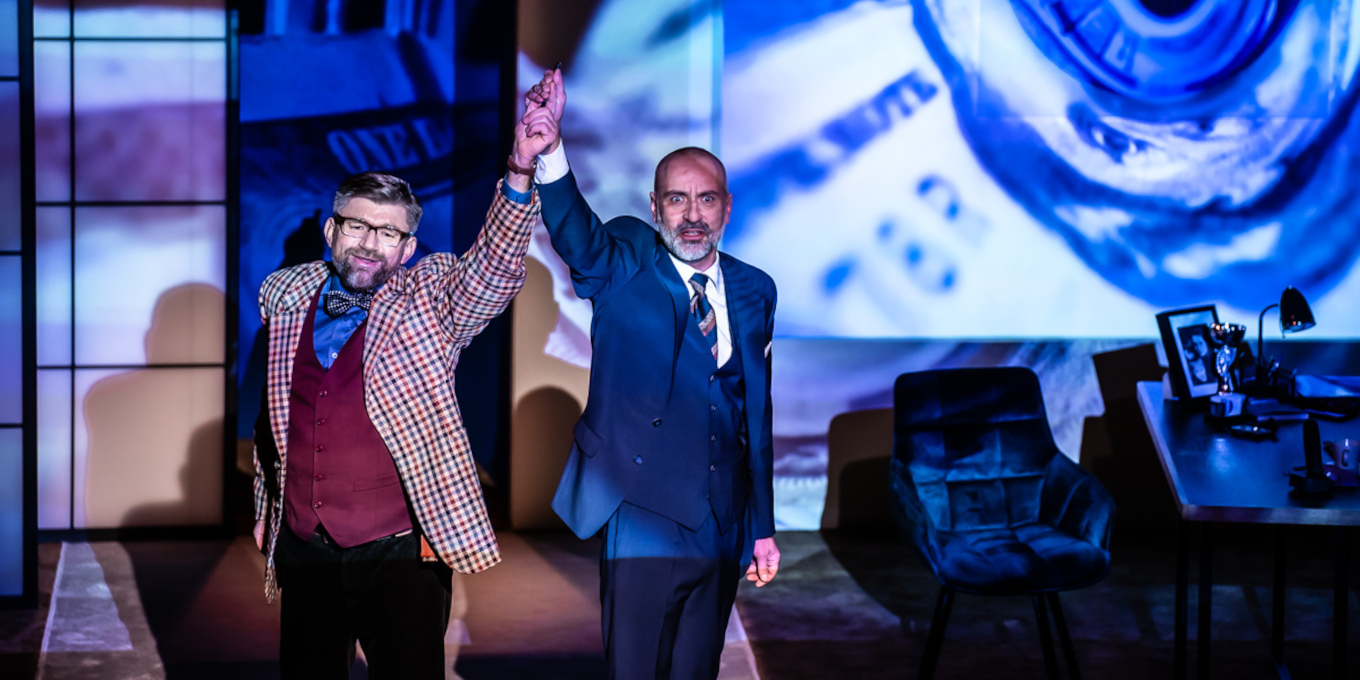 Slajd: Zdjęcie dwóch mężczyzn w garniturach. Jeden z nich unosi rękę drugiego wysoko do góry w geście wskazywania zwycięscy. Na drugim planie po prawej biurko i krzesło. Na biurku materiały biurowe, zdjęcie w ramce i lampka. W tle wizualizacje z przewagą białego i niebieskiego.