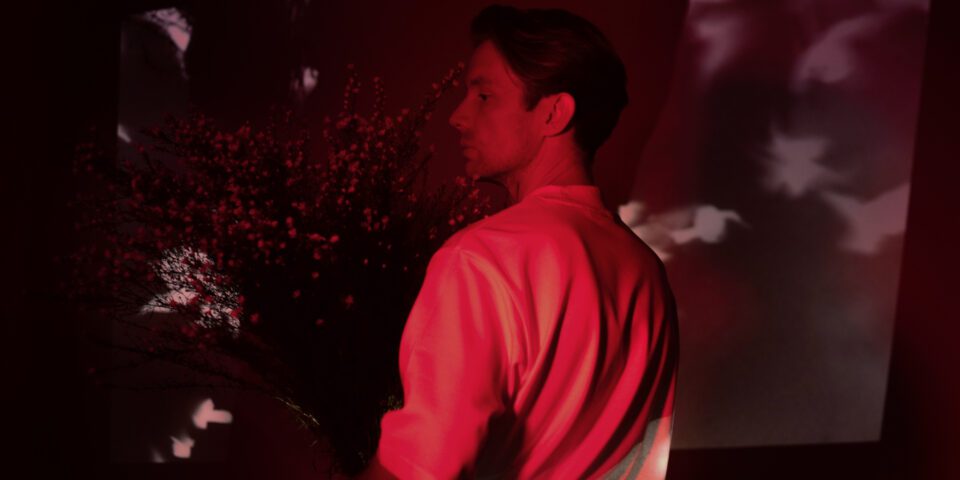 Artystyczne zdjęcie Bartka Wąsika. Jest w ujęciu z profilu, w czerwonym świetle, w którym wyróżni się jego jasna koszula na ciemnym tle. Trzyma oburącz wielki bukiet kwiatów. Ma poważną minę.