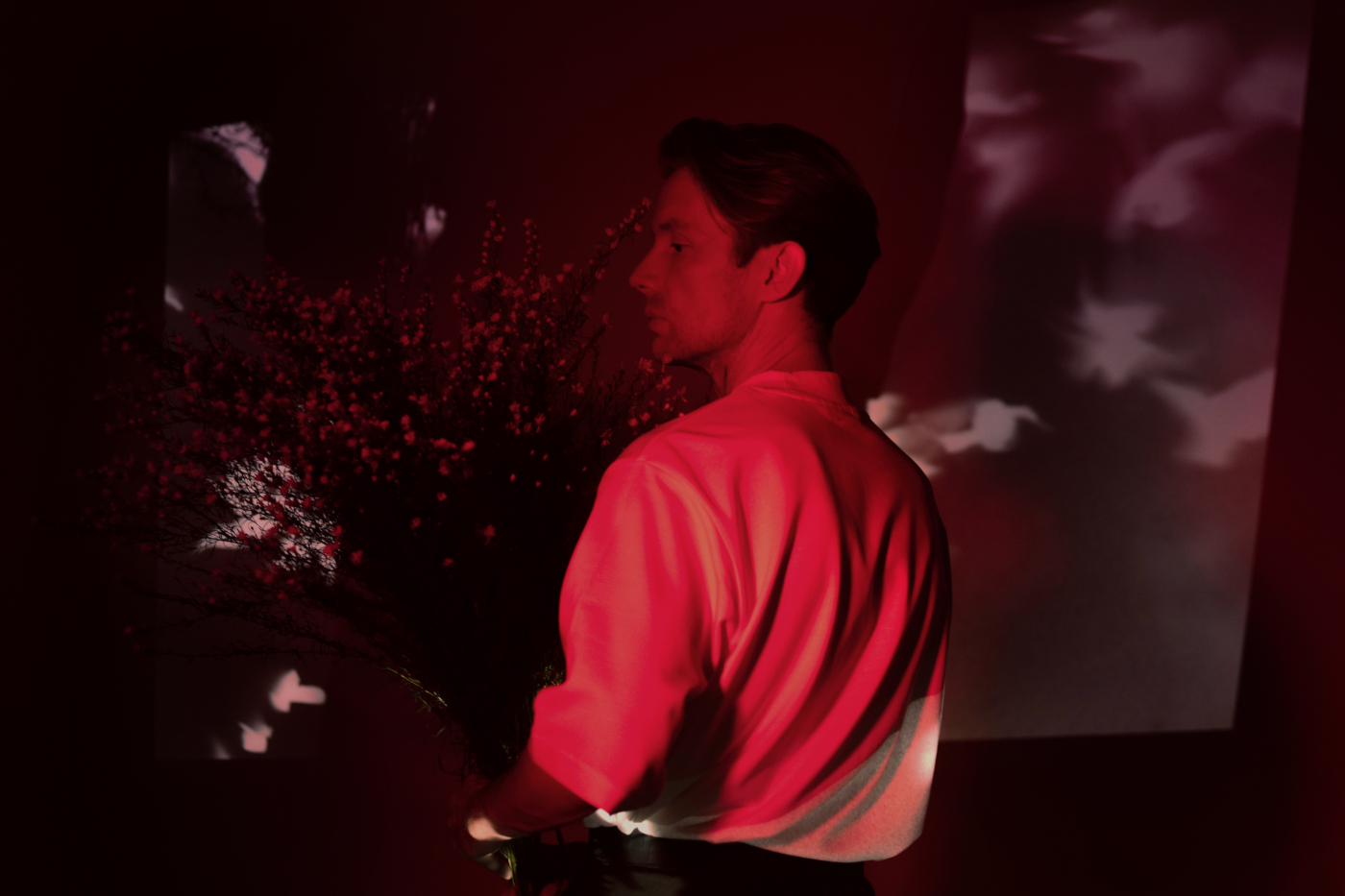 Artystyczne zdjęcie Bartka Wąsika. Jest w ujęciu z profilu, w czerwonym świetle, w którym wyróżni się jego jasna koszula na ciemnym tle. Trzyma oburącz wielki bukiet kwiatów. Ma poważną minę.