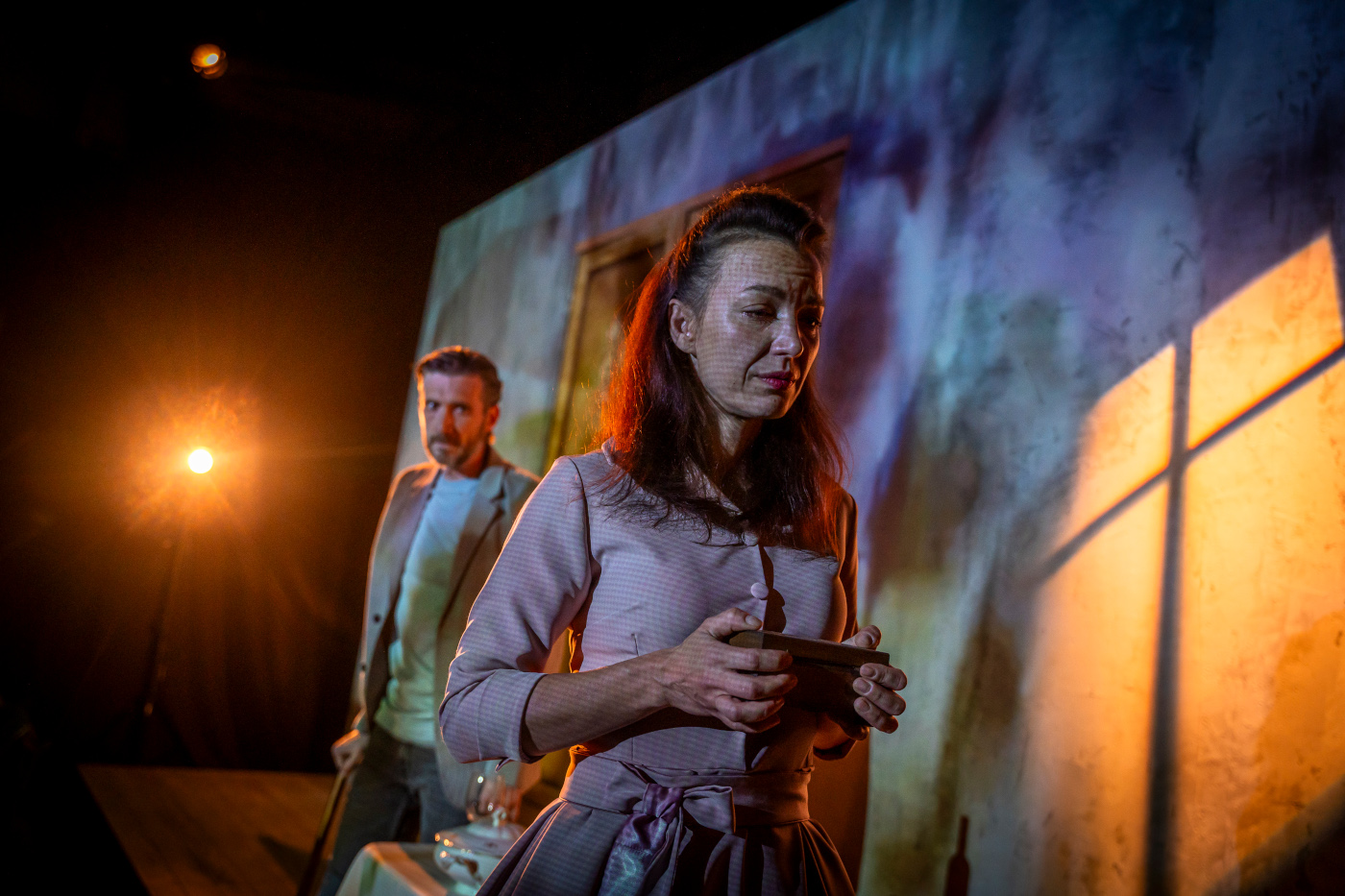 Zdjęcie kobiety i mężczyzny na scenie. Na pierwszym planie ciemnowłosa kobieta w bladoróżowej sukience. Ma smutną minę, trzyma małe, drewniane pudełko. Na drugim planie mężczyzna w jasnym stroju.