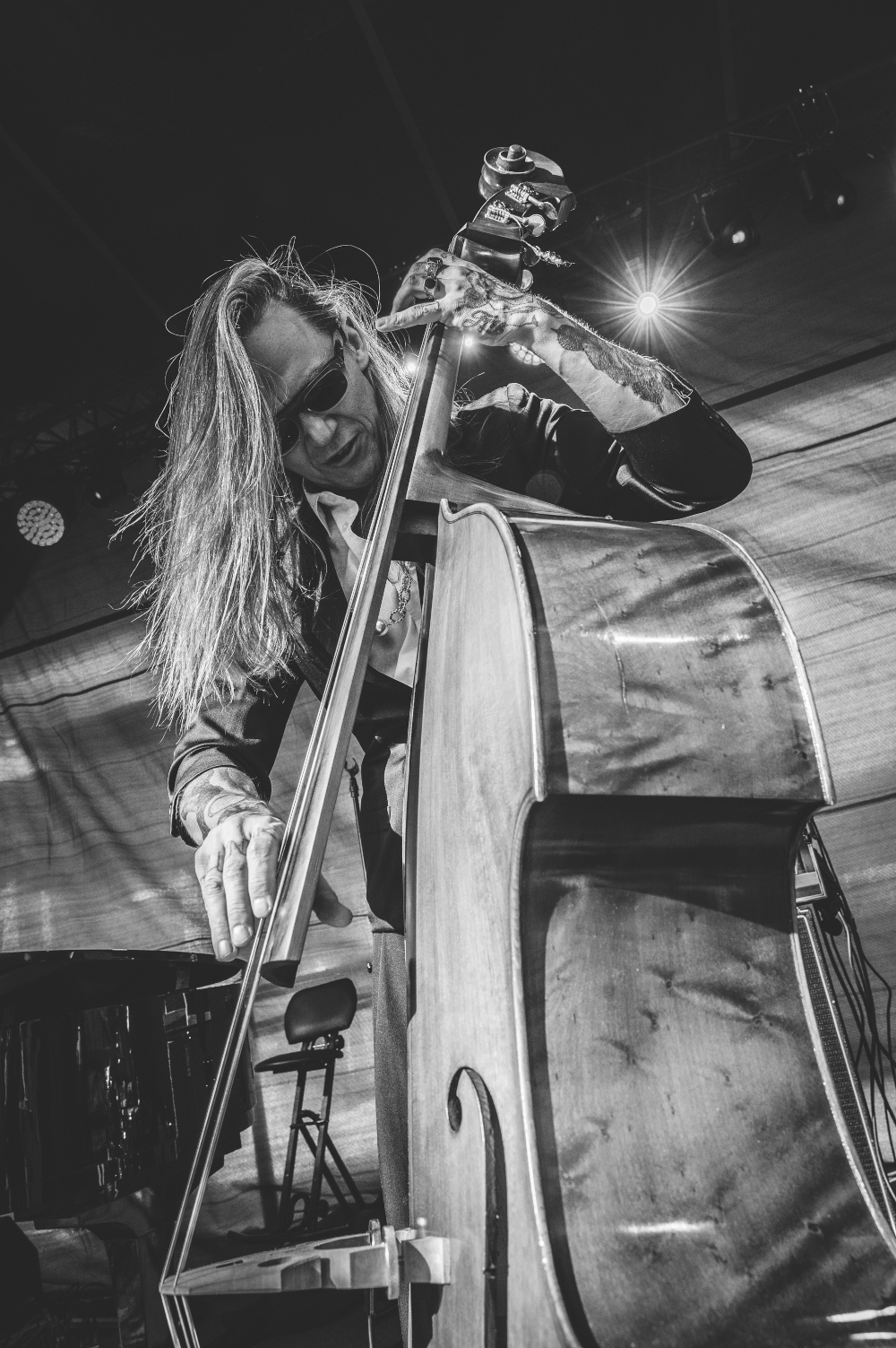 Czarno-białe zdjęcie Wojtka Mazolewskiego grającego na kontrabasie. Mężczyzna ma długie, rozpuszczone włosy i ciemne okulary, jest ubrany w białą koszulę i skórzaną kurtkę. Spod podwiniętych rękawów wystają tatuaże pokrywające całe ręce.