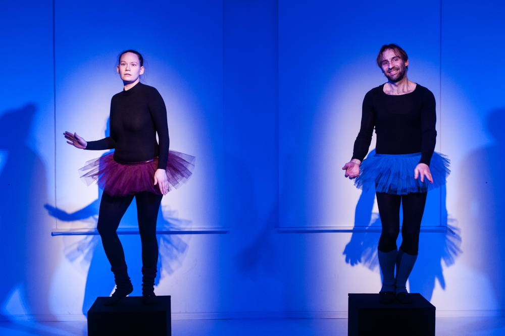 Zdjęcie kobiety i mężczyzny na niebieskim tle. Oboje stoją na sześciennych czarnych podestach. Sa ubrani w czarne dopasowane stroje, leginsy i bluzki z długimi rękawami. Każde z nich ma krótką, tiulową spódniczkę, charakterystyczną dla baletu. Spódniczka kobiety jest czerwona, a mężczyzny niebieska.