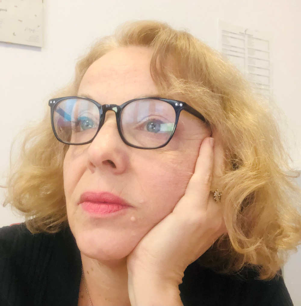 Zbliżenie na twarz kobiety w okularach. Kobieta opiera głowę na dłoni. Ma czarny strój i jasne, falowane włosy. W tle jasna ściana.