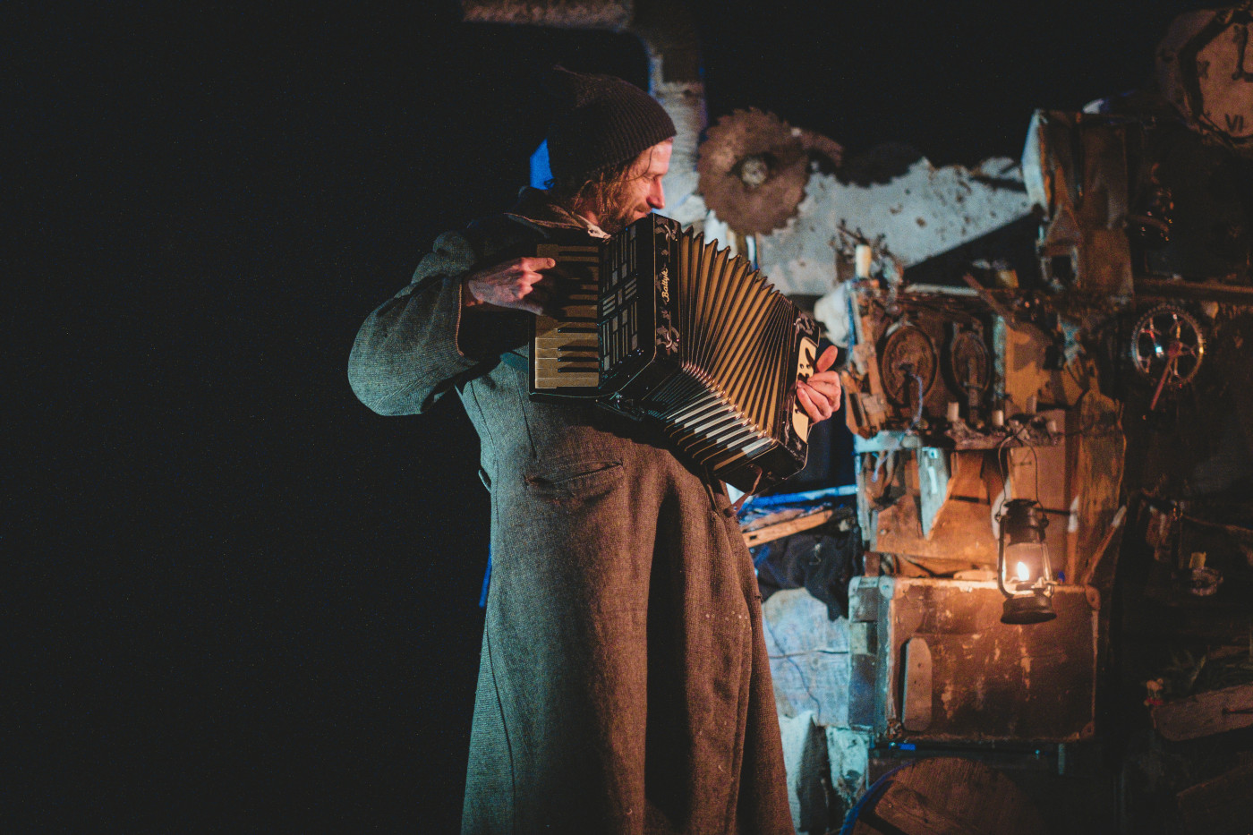 Zdjęcie przedstawia brodatego mężczyznę w średnim wieku grającego na akordeonie. Jest on ubrany w stary, brązowy płaszcz, na głowie ma czarną wełnianą czapkę spod której wystają długie, splątane włosy. Za nim znajduje się ciemne pomieszczenie wypełnione rupieciami, z prawej strony oświetlone światłem lampy naftowej.