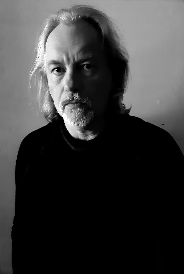 Czarno-białe zdjęcie portretowe mężczyzny. Ma jasne, długie włosy, krótką brodę i wąsy. Jest w czarnej bluzie. Tło jasnoszare.