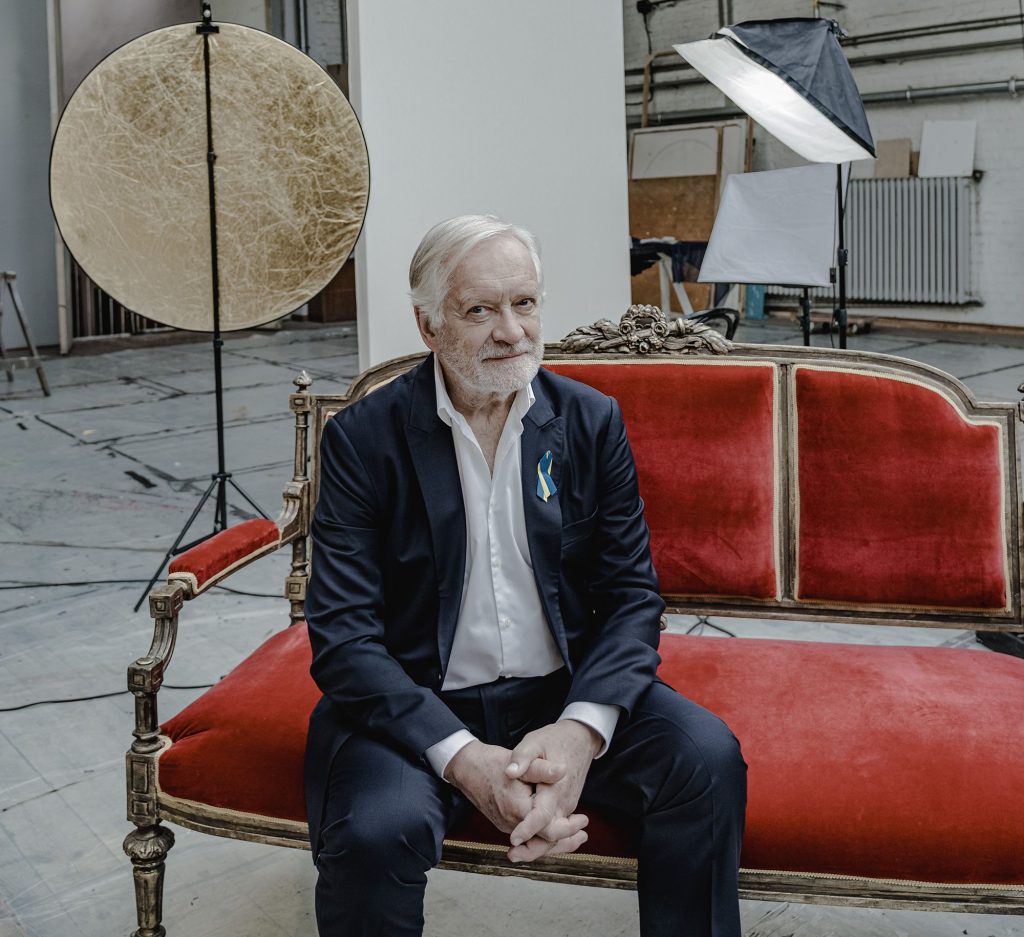 Zdjęcie Andrzeja Seweryna. Mężczyzna siedzi na czerwonej sofie na drewnianych nóżkach. Jest ubrany w białą koszulę i ciemny garnitur. W tle duże pomieszczenie z kilkoma rekwizytami ze studia fotograficznego, reflektorem i złotą, okrągłą blendą.