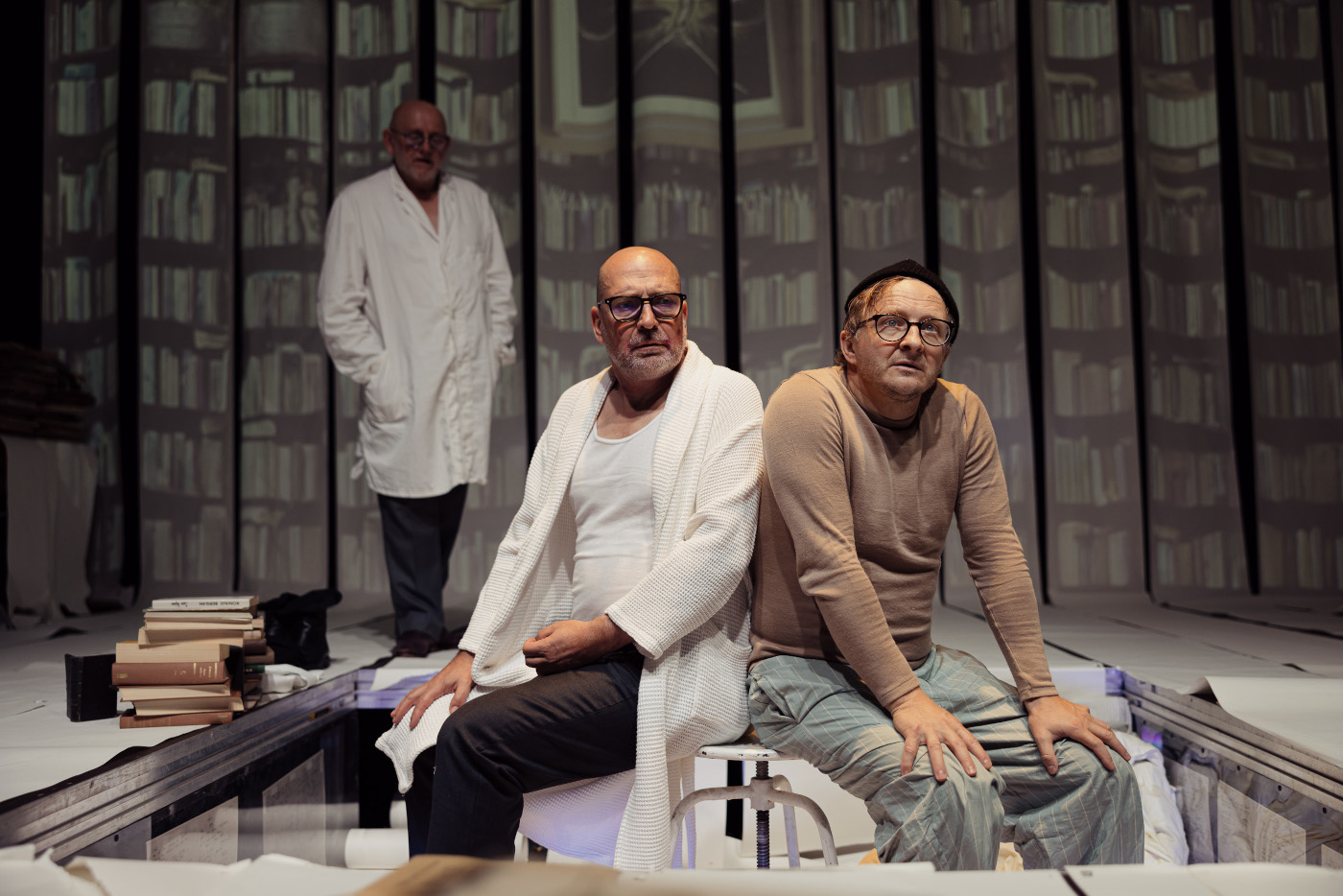 Zdjęcie trzech mężczyzn na scenie. Na pierwszym planie dwóch mężczyzn siedzących na białym stołku. Jeden z nich w białym fartuchu. Na drugim planie po lewej stoi trzeci mężczyzna w białym fartuchu. W tle wizualizacja ze ścianą wypełnioną półkami z książkami.