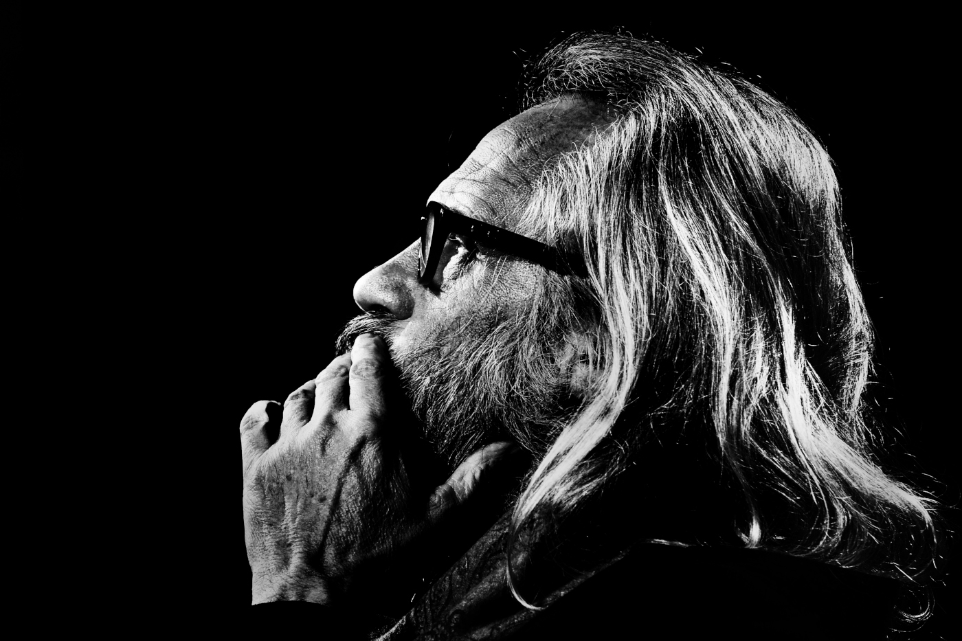 Czarno-białe zdjęcie Janusza Opryńskiego. Zbliżenie na twarz i dłoń zasłaniającą podbródek. Mężczyzna ma długie, jasne włosy, zarost i okulary w grubych, ciemnych oprawkach. Tło jest czarne.