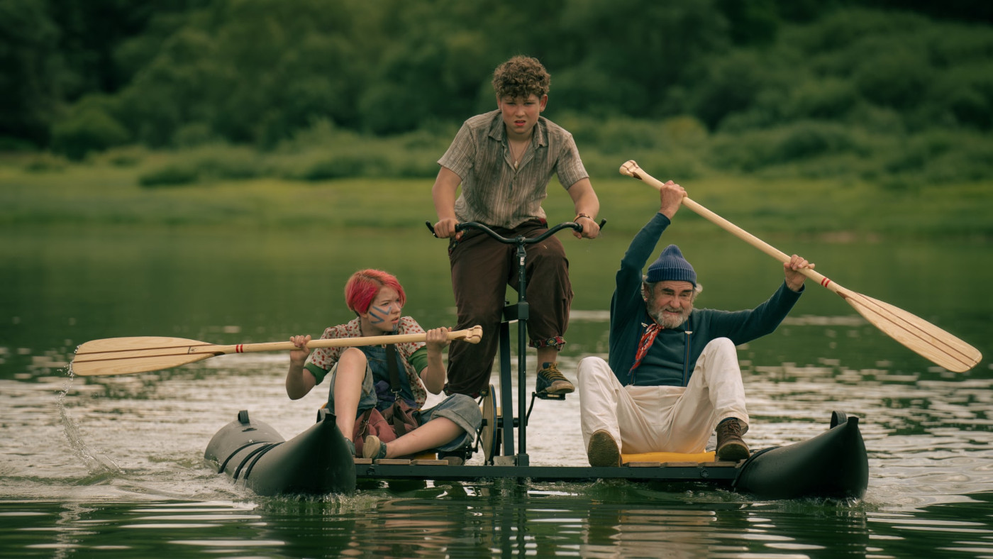 Zdjęcie trzech osób na konstrukcji pływającej po jeziorze. W centrum symetrycznej konstrukcji jest rower, na którym jest chłopiec. Po bokach są podłużne pływaki, a pomiędzy pływakami a rowerem siedzenia. Po lewej siedzi dziewczynka z rudymi włosami. Po prawej starszy mężczyzna. Oboje trzymają wiosła. W tle u dołu tafla jeziora, u góry las.