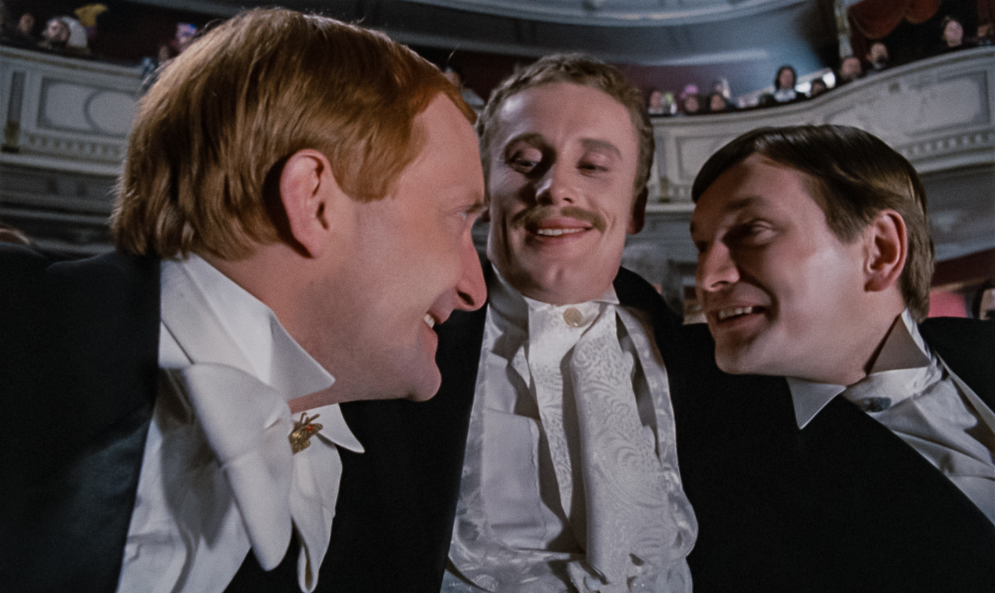 Trzech uśmiechniętych mężczyzn w eleganckich, wieczorowych strojach. W tle wnętrze teatru z lożami na balkonie.