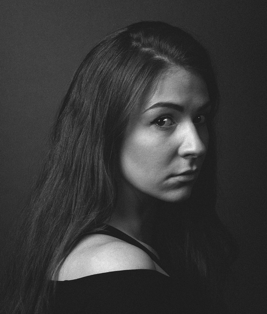 Czarno-białe zdjęcie portretowe kobiety w ujęciu z półprofilu. Ma długie, ciemne włosy i ciemny strój. Tło jest ciemnoszare.
