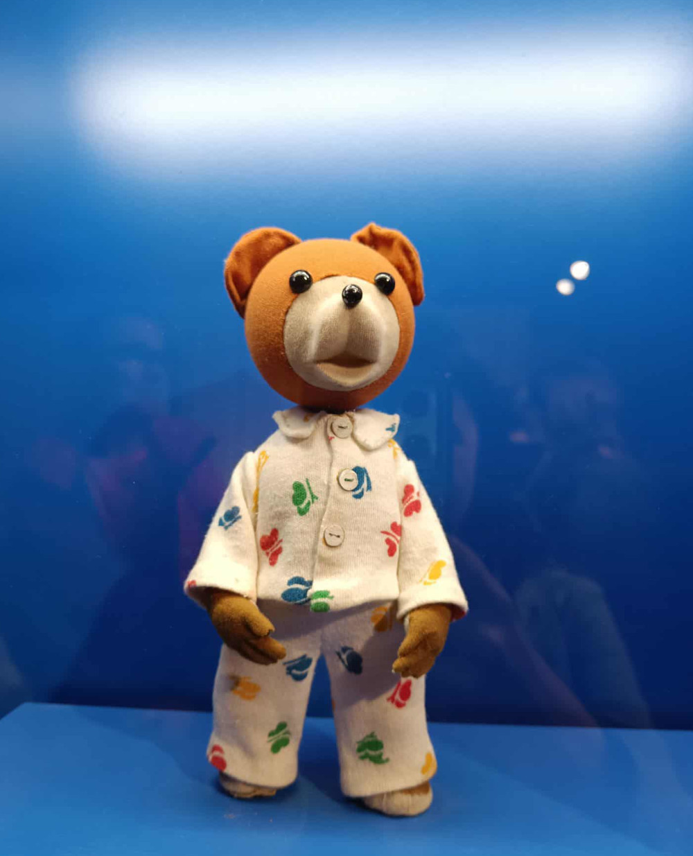 Zdjęcie misia uszatka w pozycji stojącej. Jest ubrany w białą piżamę z kolorowym deseniem. Tło niebieskie.