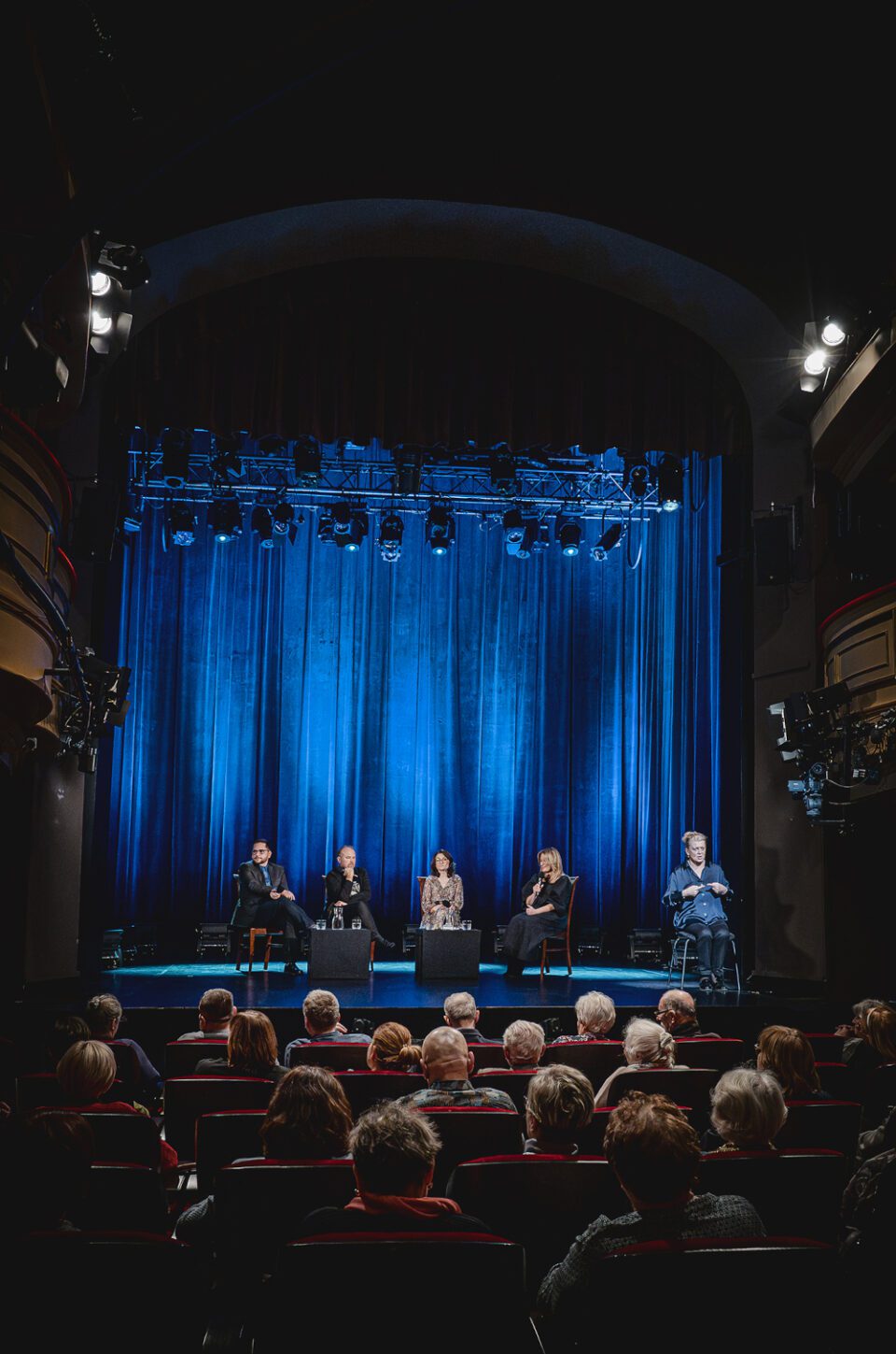 Zdjęcie sceny zza pleców widzów siedzących w rzędach. Na scenie pięć osób. Cztery osoby siedzą przy stoliku mieszczonym w centrum. Piąta osoba to tłumaczka na polski język migowy siedząca skrajnie po prawej stronie. W tle kotara oświetlona na niebiesko.