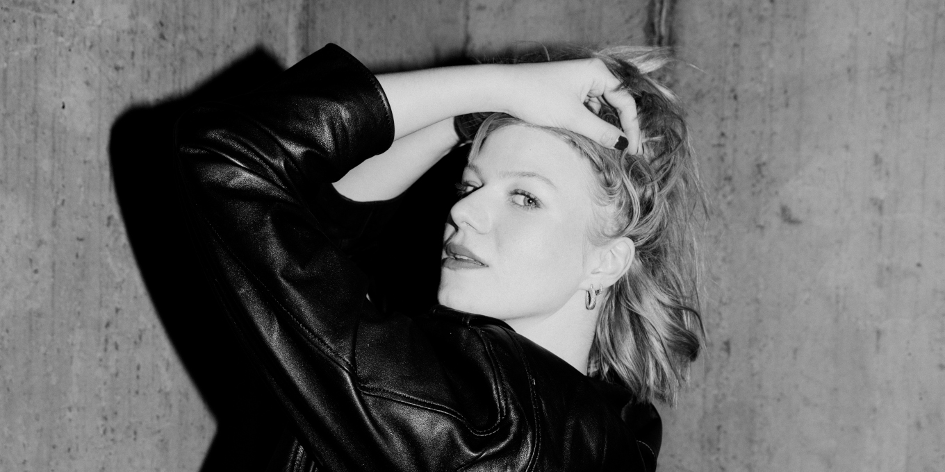 Slajd: Czarno-białe zdjęcie młodej kobiety w ujęciu z profilu. Kobieta dotyka rękami swoich włosów. Jest ubrana w skórzaną kurtkę. Tło szare.