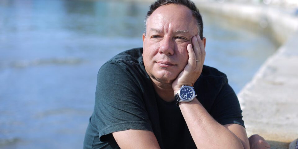 Zdjęcie mężczyzny w czarnej koszulce z krótkim rękawem. Mężczyzna opiera twarz na dłoni. W rozmytym tle krajobraz ze zbiornikiem wodnym.