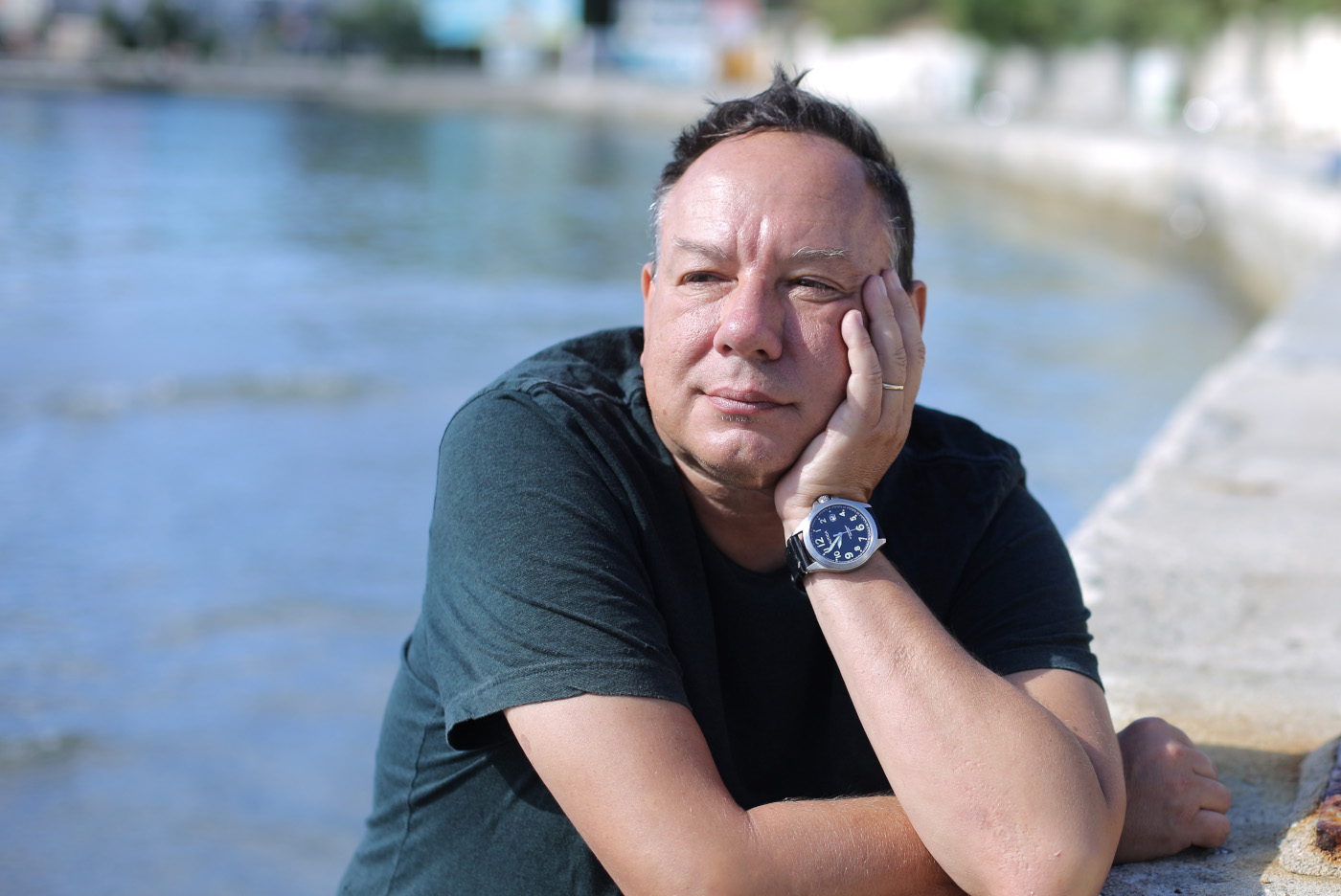 Zdjęcie mężczyzny w czarnej koszulce z krótkim rękawem. Mężczyzna opiera twarz na dłoni. W rozmytym tle krajobraz ze zbiornikiem wodnym.