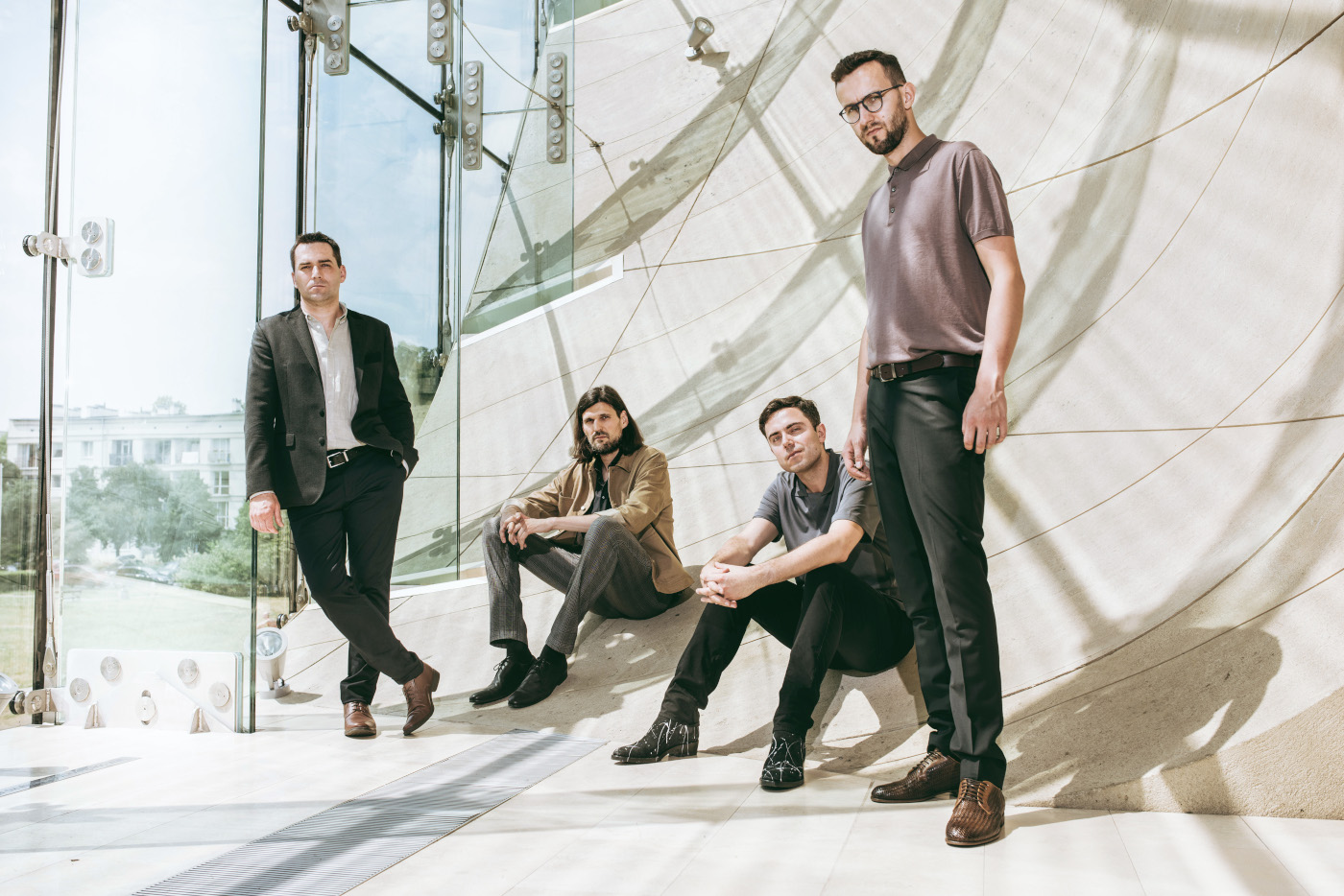 Zdjęcie trzech mężczyzn. Dwóch po środku siedzi, panowie po bokach stoją. Wszyscy mają proste stroje w ciemnych stonowanych kolorach. Znajdują się w nowoczesnej przestrzeni z dużymi przeszkleniami, za którymi widać zieleń i błękit. Podłoga i ściana są wyłożone jasnym kamieniem.