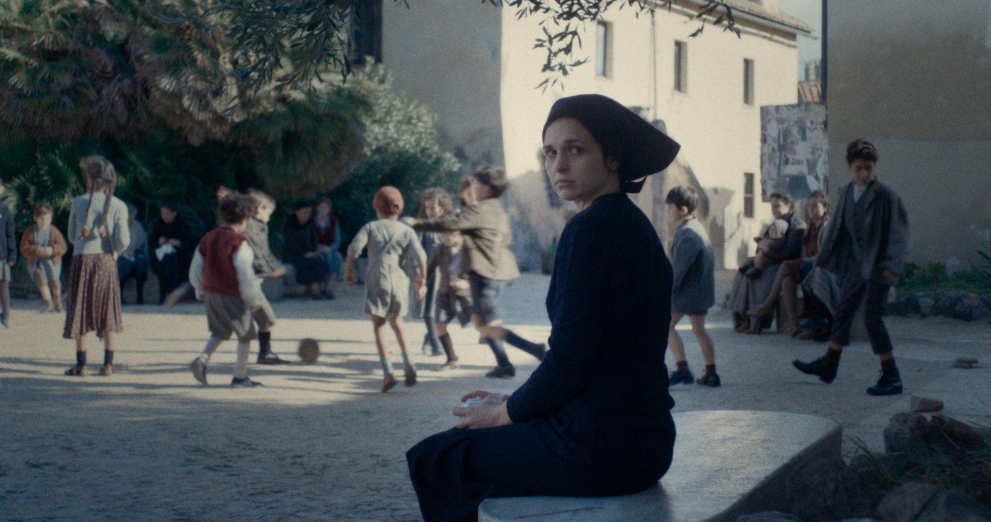 Na pierwszym planie smutna kobieta w czarnym stroju z czarną chustką na włosach. Siedzi na kamiennej ławce. Na drugim planie plac na którym kilkanaścioro dzieci gra w piłkę. Mają skromne stroje, charakterystyczne dla pierwszej połowy dwudziestego wieku. W tle po lewej drzewa, po prawej jasne budynki.