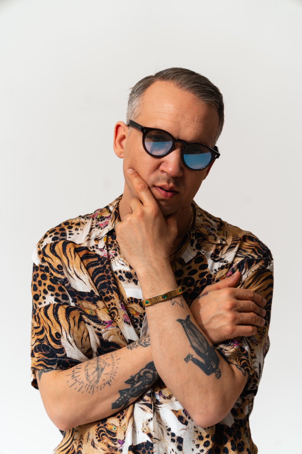 Zdjęcie portretowe Jana Młynarskiego. Jest ubrany w koszulę w tygrysy i lamparty. Ma krótkie włosy i ciemne okulary. Ma odsłonięte przedramiona i tatuaże. Ma skrzyżowane ręce. Jedną dłonią dotyka przeciwległego przedramienia, drugą trzyma na podbródku. Tło jest białe.