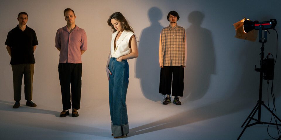 Zdjęcie czterech osób, jednej kobiety i trzech mężczyzn. Stoją w odstępach. Kobieta najbardziej z przodu. Po prawej stronie stoi reflektor na statywie. Postacie rzucają cienie na podłogę i ścianę.
