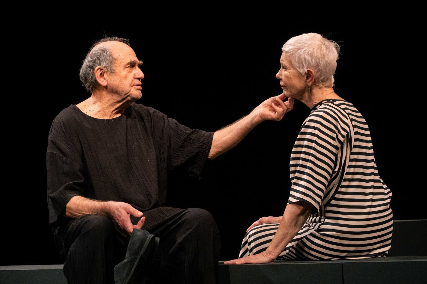 Zdjęcie kobiety i mężczyzny, którzy siedzą naprzeciwko siebie. Mężczyzna po lewej, kobieta po prawej. Mężczyzna ma czarny, luźny strój. w jednej ręce trzyma czarne buty, drugą dłonią dotyka delikatnie podbródka kobiety. Kobieta ma luźną sukienkę w czarno-białe poprzeczne paski, ma krótkie, białe włosy, dłonie opiera na siedzisku. Tło jest czarne. Kobieta i mężczyzna to postacie grane w spektaklu przez Jana Peszka i Małgorzatę Zajączkowską.