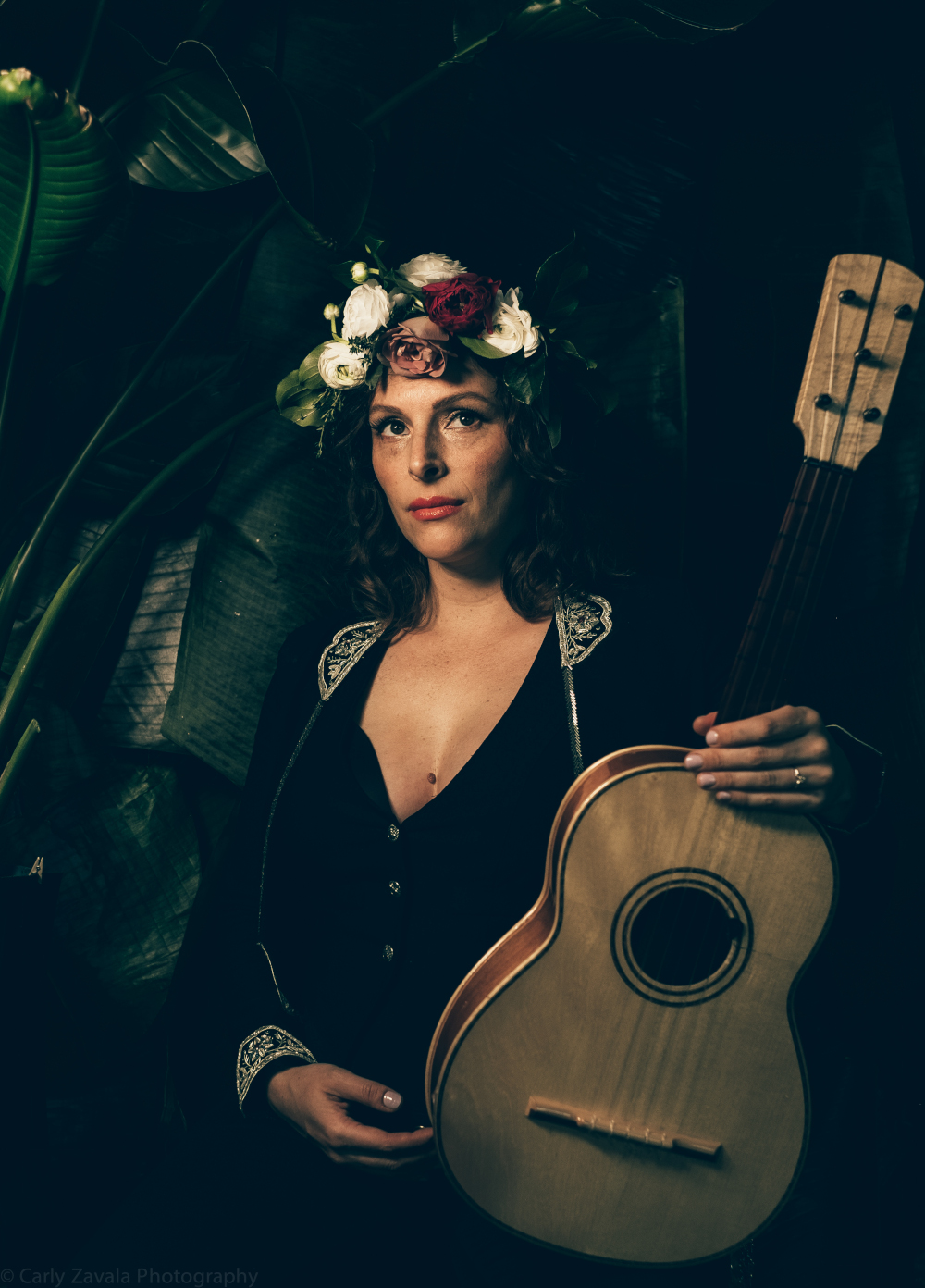 Zdjęcie kobiety z gitarą akustyczną. Kobieta ma czarny strój ze srebrnymi naramiennikami i mankietami. Ma ciemne włosy i wianek z kwiatów na głowie. Tło jest ciemne.