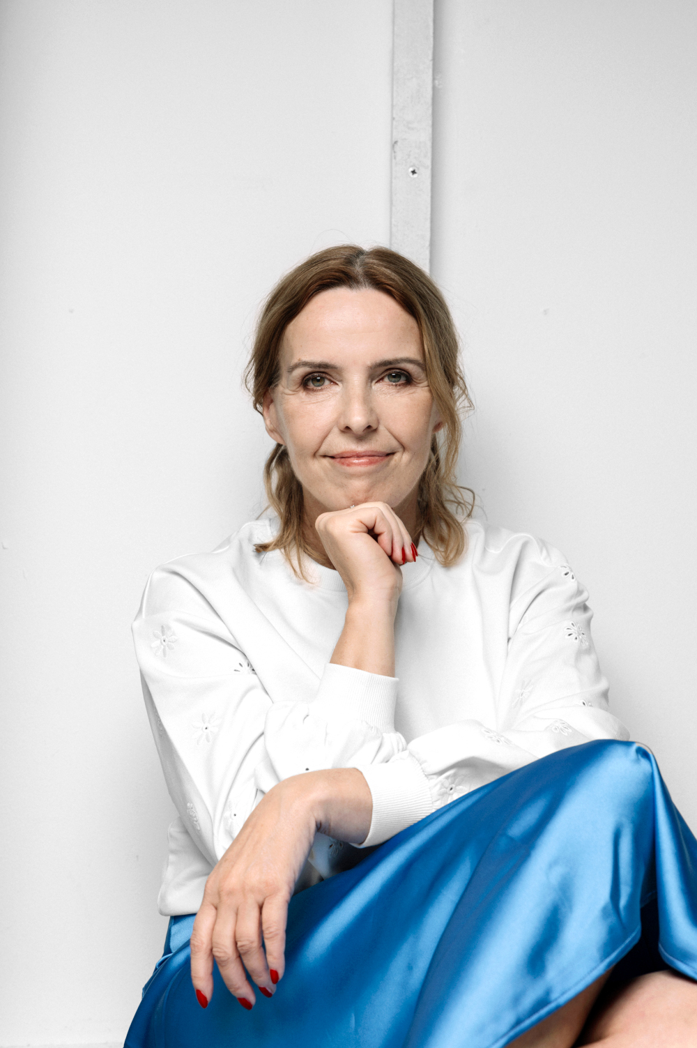 Zdjęcie uśmiechniętej kobiety w pozycji siedzącej. Ma upięte po bokach włosy. Jedną dłonią podpiera podbródek. Jest ubrana w białą koszulę i niebieską, atłasową spódnicę. Tło jest białe.